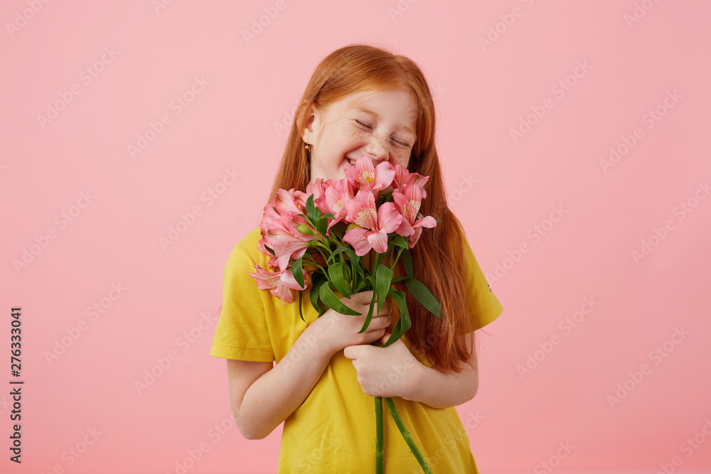 Portrait petite smiles freckles red-haired girl with two tails, looks cute, wears in yellow t-shirt, holds bouquet and stands over pink background, enjoying the smell of flowers.
