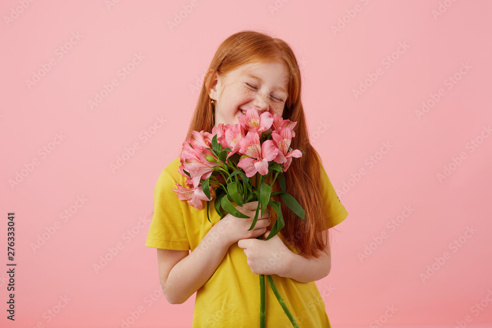 Portrait petite smiles freckles red-haired girl with two tails, looks cute, wears in yellow t-shirt, holds bouquet and stands over pink background, enjoying the smell of flowers. Portrait petite smiles freckles red-haired girl with two tails, looks cute, wears in yellow t-shirt, holds bouquet and stands over pink background, enjoying the smell of flowers.