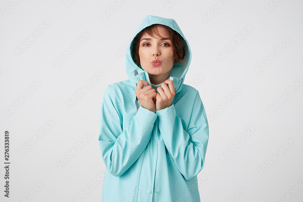 Portrait of positive young nice lady in blue rain coat, with a hood on his head, looks at the camera with happy expressions, send kiss at the camera, stands over white wall. Portrait of positive young nice lady in blue rain coat, with a hood on his head, looks at the camera with happy expressions, send kiss at the camera, stands over white wall.