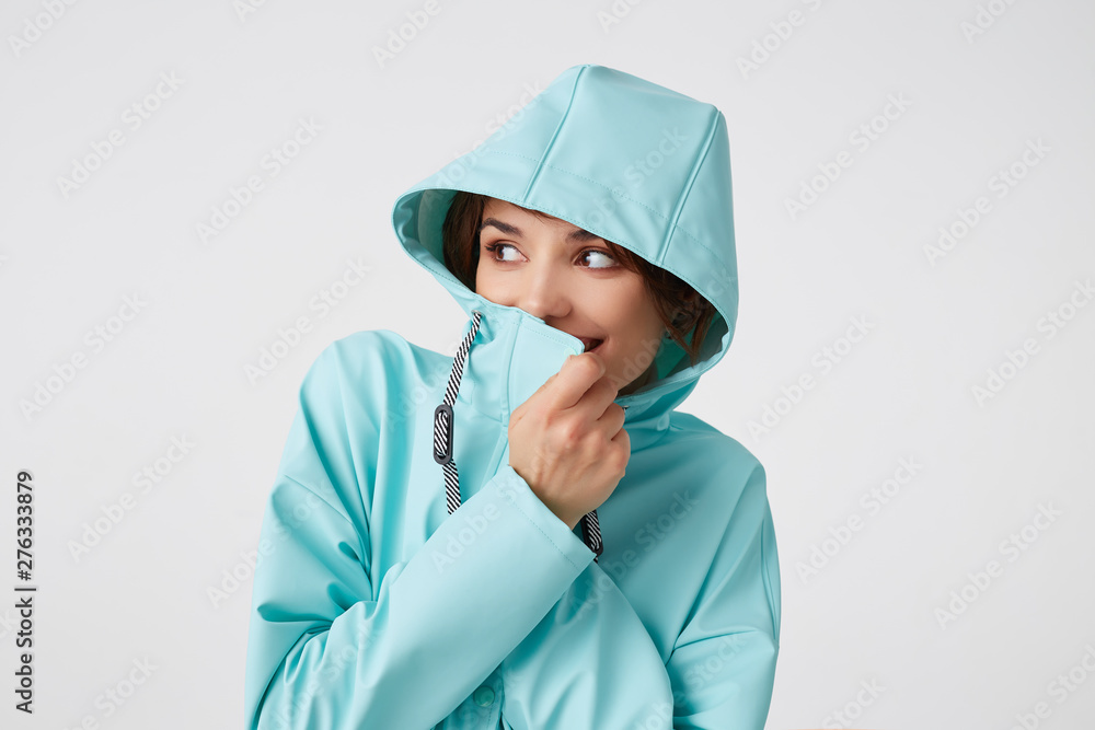 Portrait of positive young nice lady in blue rain coat, with a hood on his head, looks away with happy expressions, broadly smiling, covers his face with a collar, stands over white wall. Portrait of positive young nice lady in blue rain coat, with a hood on his head, looks away with happy expressions, broadly smiling, covers his face with a collar, stands over white wall.