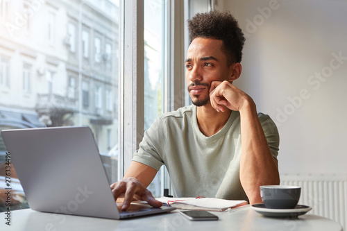 Принти на полотні Portrait of young attractive African American thinking boy, sits at a table in a cafe, works at a laptop and drinks aromatic coffee and thoughtfully looks at the window