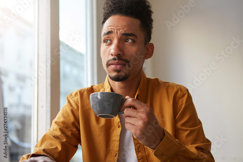 Portrait of young handsome dark skinned thinking man, drinks aromatic coffee from a gray cam and thoughtfully looks away. Maby he wonts to be barista? photo