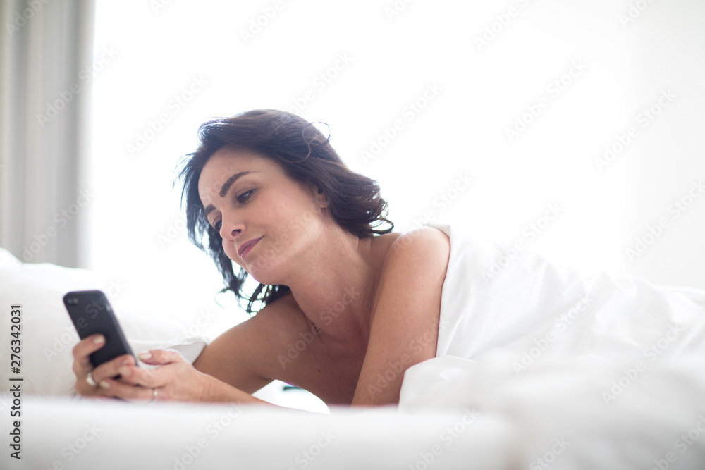 Beautiful young woman using her smart phone in bed, following social media, being quite addicted to this device without realizing Beautiful young woman using her smart phone in bed, following social media, being quite addicted to this device without realizing