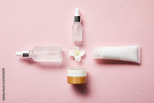 flat lay of cosmetic glass bottles, jar, cream tube with cream and jasmine flower in middle on pink