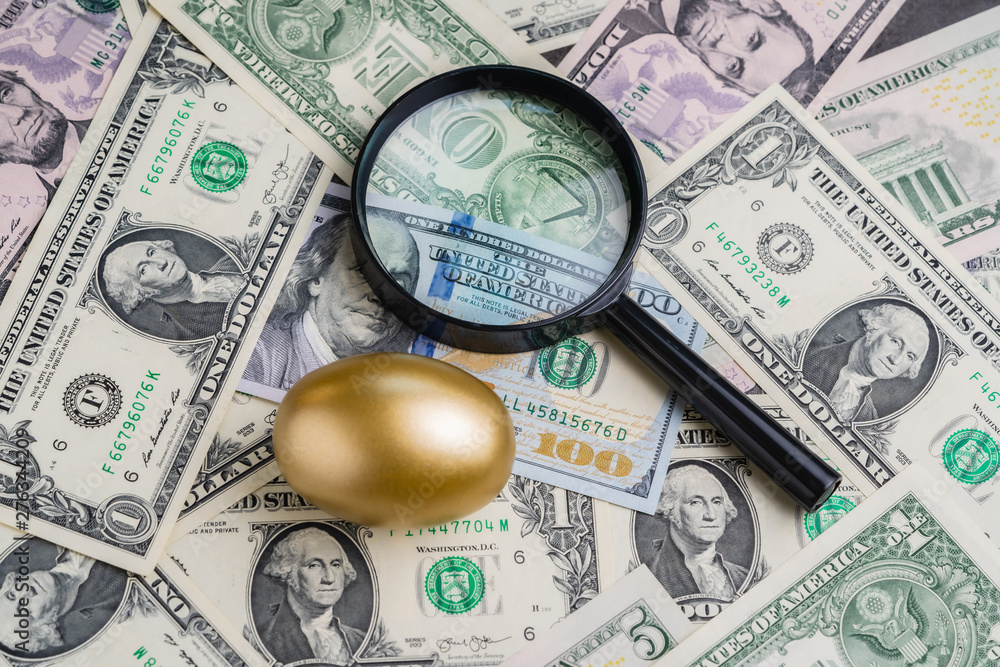 Magnifying glass with shiny golden egg under pile of US America dollar banknotes money metaphor of precious wealth, finding good stock with high dividend or success investment in stock market concept Magnifying glass with shiny golden egg under pile of US America dollar banknotes money metaphor of precious wealth, finding good stock with high dividend or success investment in stock market concept