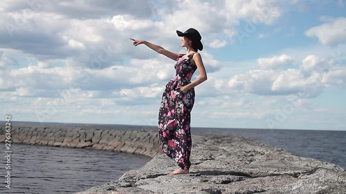 A young beautiful girl in a long dress and a black hat standing barefoot on a dam near the water shows her hand to the side and smiles