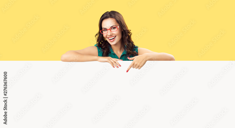 Happy smiling beautiful young woman in green clothing and red glasses, pointing blank white bill board or copy space empty place for ad slogan or sign text, over yellow color background Happy smiling beautiful young woman in green clothing and red glasses, pointing blank white bill board or copy space empty place for ad slogan or sign text, over yellow color background