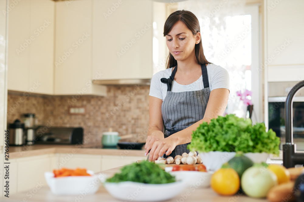 Beautiful smiling dedicated Caucasian brunette in apron standing in kitchen and chopping mushrooms. On table are lots of vegetables. Cooking at home concept.