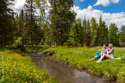 A picnic on the bank of a mountain river with green grass and yellow flowers against the background of coniferous trees and a blue sky with clouds; a beautiful blonde is sitting with man