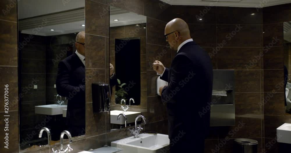 In the men's room, a bald man in glasses and a business suit, standing near the sink, threatens himself in the mirror with his fist, washes his hands, then takes a paper towel and wipes their hands In the men's room, a bald man in glasses and a business suit, standing near the sink, threatens himself in the mirror with his fist, washes his hands, then takes a paper towel and wipes their hands