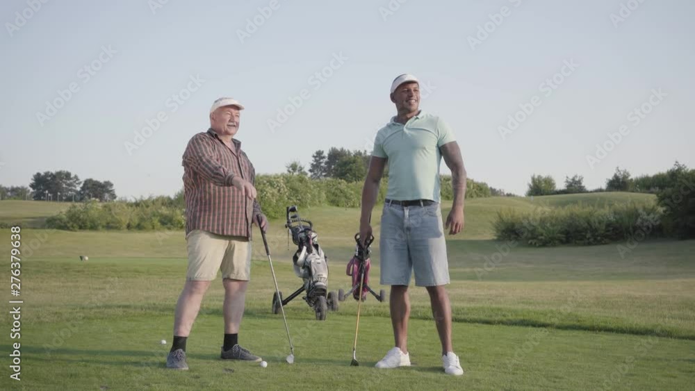 Mature Caucasian man and young middle eastern man playing golf on the golf course. Men gesture to the side, look and discuss. Summer leisure. Mature Caucasian man and young middle eastern man playing golf on the golf course. Men gesture to the side, look and discuss. Summer leisure.