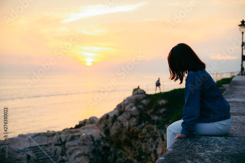 young pretty woman sitting at cliff looking on beautiful sunset © phpetrunina14