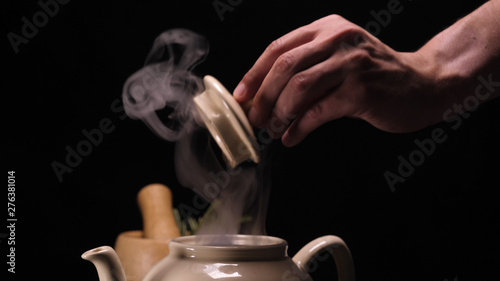On a black background, a brewed teapot (teapot) on a tray is a lemon and a grassy saucer, from the kettle a lot of steam comes out revealing a crush, hot water is filled and tea in the saucer in a mug