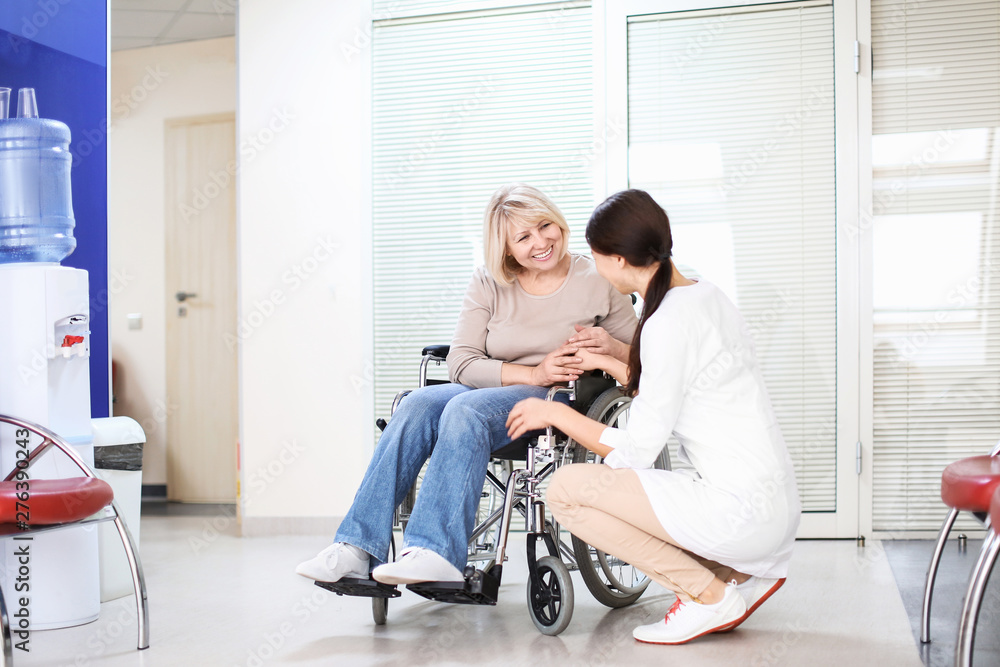 Young female doctor taking care of mature woman in wheelchair indoors