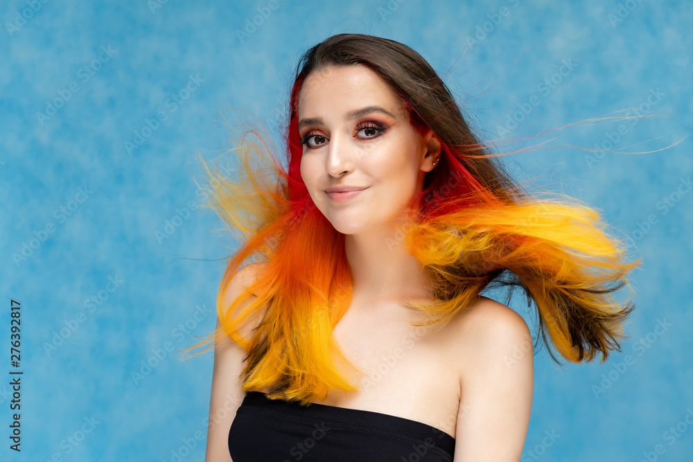 A close-up portrait photo of a fashionable hairstyle red-yellow in studio on a blue background. The pretty brunette model with beautiful make-up has beautiful flowing colorful hair. A close-up portrait photo of a fashionable hairstyle red-yellow in studio on a blue background. The pretty brunette model with beautiful make-up has beautiful flowing colorful hair.