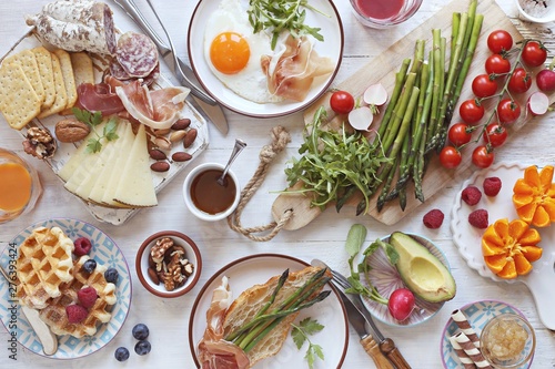  Brunch or breakfast table, meal variety with fried egg, asparagus, avocado, sausage and cheese variety, croissants, smoothie, fresh waffles and fruits. Overhead view