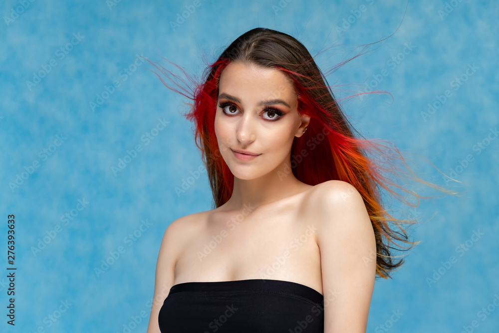 A close-up portrait photo of a fashionable hairstyle red-yellow in studio on a blue background. The pretty brunette model with beautiful make-up has beautiful flowing colorful hair.