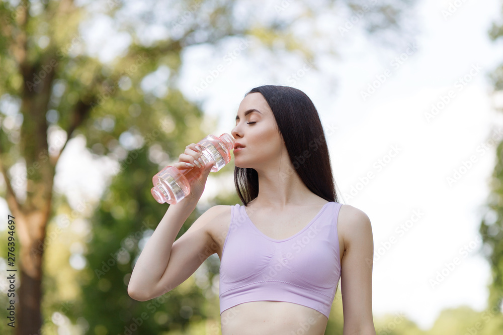 Beautiful slim brunette woman drinking water from bottle after running at the morning park to stay hydrated. Female fitness model working out outdoor. Concept of healthy lifestyle. Beautiful slim brunette woman drinking water from bottle after running at the morning park to stay hydrated. Female fitness model working out outdoor. Concept of healthy lifestyle.