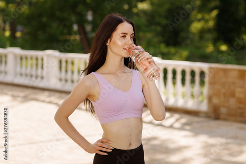 Beautiful slim brunette woman drinking water from bottle after running at the morning park to stay hydrated. Female fitness model working out outdoor. Concept of healthy lifestyle.