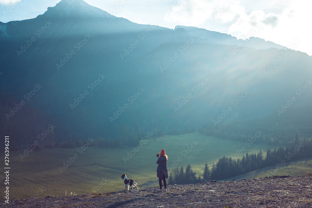 A woman with orange winter hat and Springer Spaniel dog look at Tatra mountains. Sunset landscape.