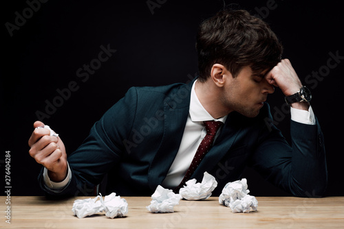 depressed businessman sitting at desk near crumpled paper isolated on black photo