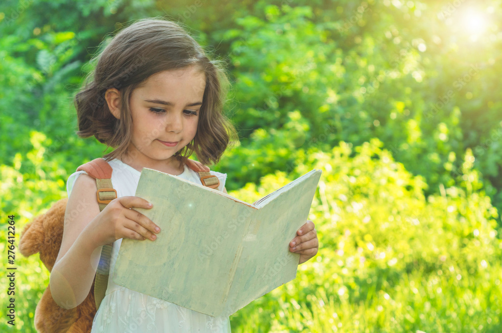 Cute industrious child is standing with a book and a briefcase outdoors. Little Girl reading the book. Place for text Cute industrious child is standing with a book and a briefcase outdoors. Little Girl reading the book. Place for text