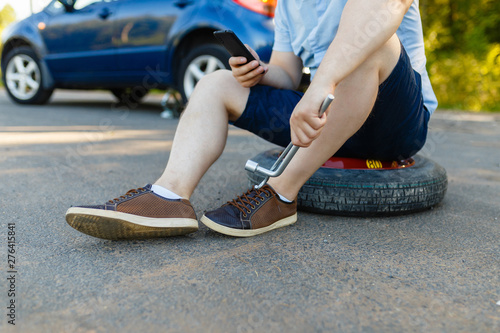 Sad and depressed person sits on a spare wheel near a blue car with a punctured tire and an open trunk. A man calls using a smartphone mobile tire service.