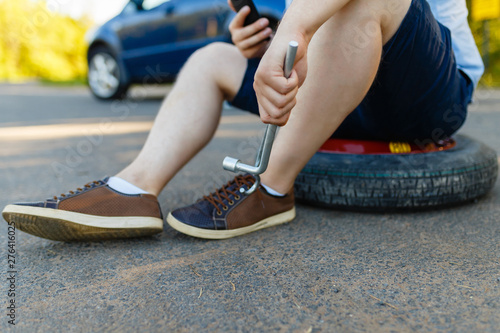 Fototapeta Naklejka Na Ścianę i Meble -  Sad and depressed person sits on a spare wheel near a blue car with a punctured tire and an open trunk. A man calls using a smartphone mobile tire service.