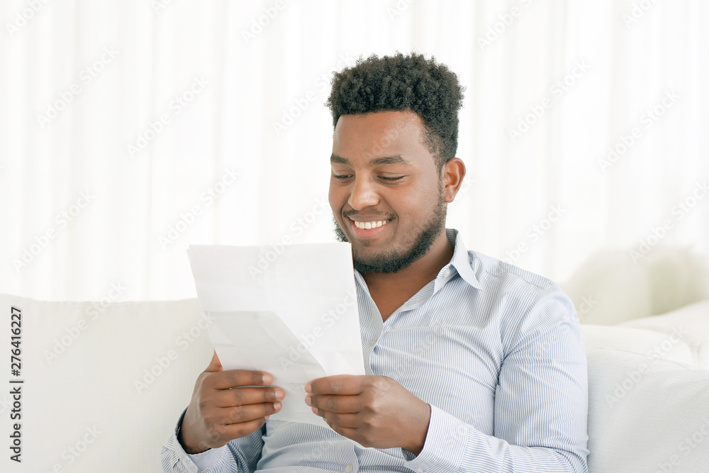 African male reads a letter with good news sitting on the sofa at home. An euphoric male is happy after reading good news in a written letter, approving a loan. African male reads a letter with good news sitting on the sofa at home. An euphoric male is happy after reading good news in a written letter, approving a loan.