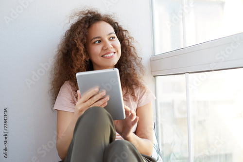 Positive young nice african american woman with curly hair sitting by the window, holding digital tablet and looks away at the window.