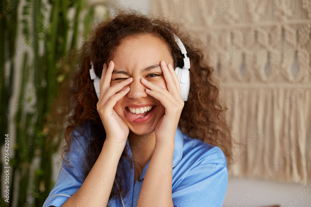 Funny curly young young woman, broadly smiles and listening favorite song in headphones, enjoying the music and Sundays morning, looks through his fingers and shows tongue.