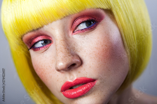 Beauty portrait of cute young female model with freckles, red makeup and yellow wig, looking away with serious face. indoor studio shot, isolated on gray background.