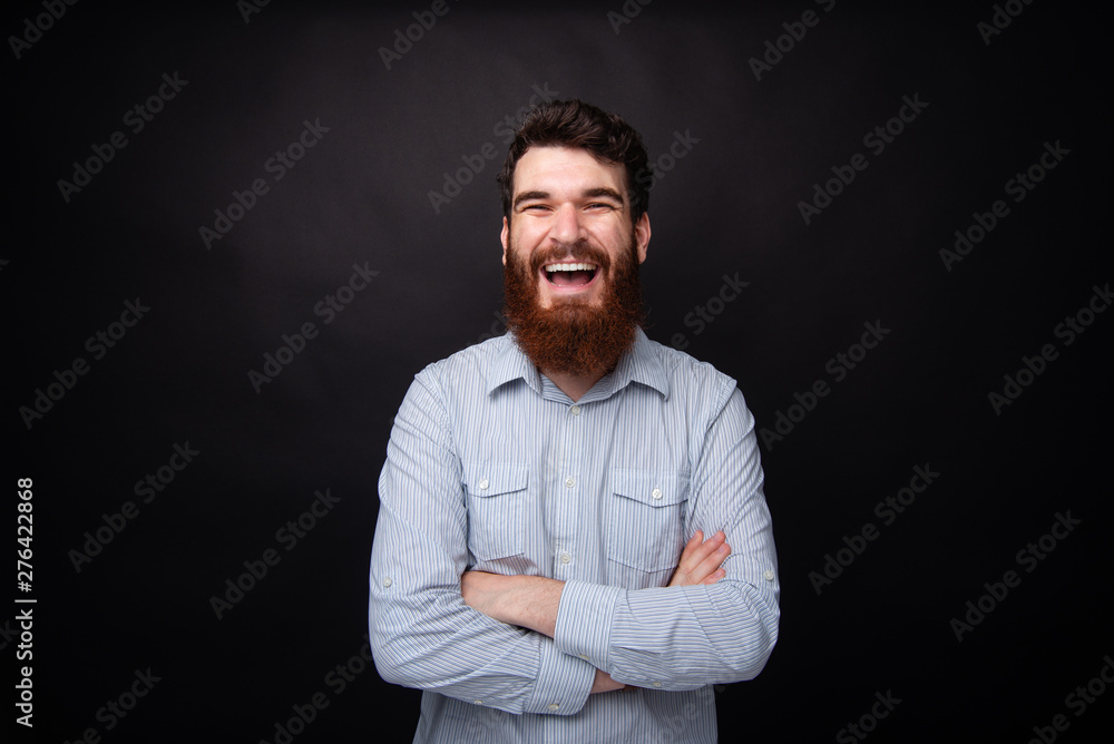 Happiness and laughing. Portrait of a young bearded man standing with arms crossed on black background laughing Happiness and laughing. Portrait of a young bearded man standing with arms crossed on black background laughing