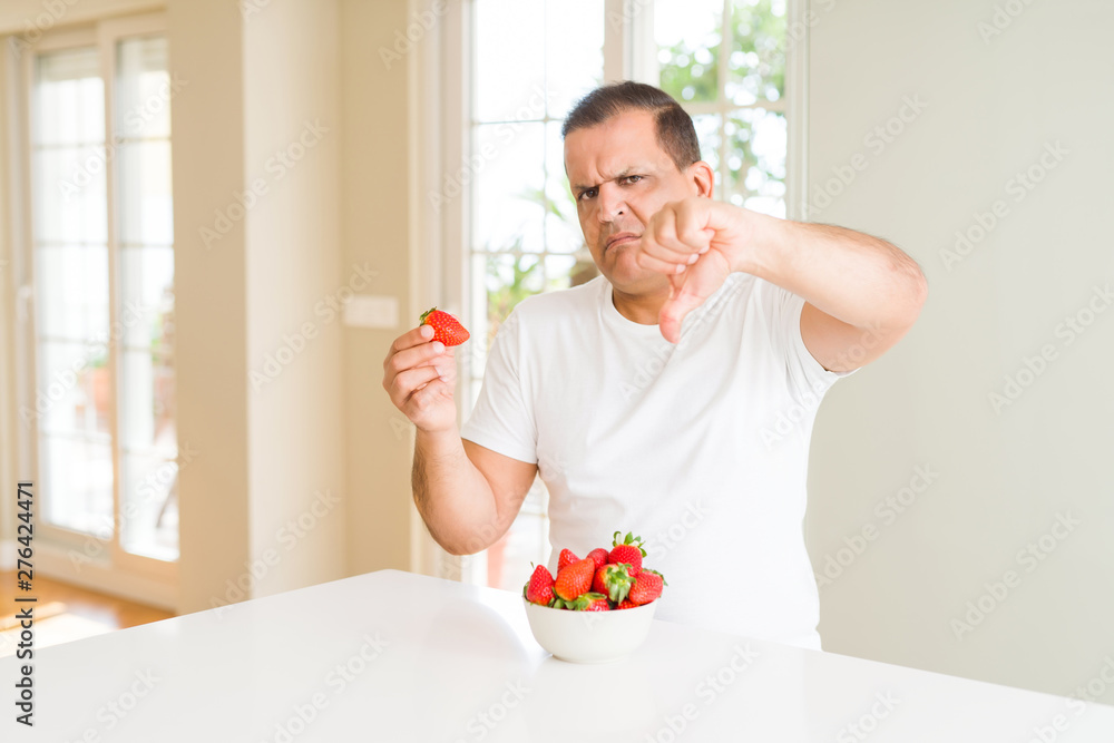 Middle age man eating strawberries at home with angry face, negative sign showing dislike with thumbs down, rejection concept Middle age man eating strawberries at home with angry face, negative sign showing dislike with thumbs down, rejection concept