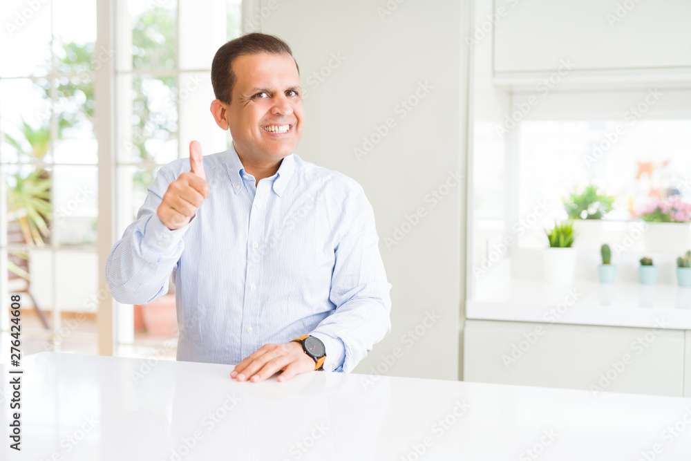 Middle age man sitting at home doing happy thumbs up gesture with hand. Approving expression looking at the camera showing success.