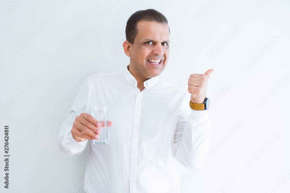 Middle age man drinking a glass of water over white background pointing and showing with thumb up to the side with happy face smiling Middle age man drinking a glass of water over white background pointing and showing with thumb up to the side with happy face smiling