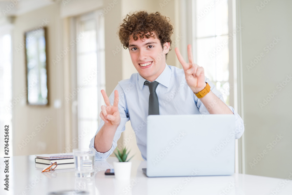 Young business man working with computer laptop at the office smiling looking to the camera showing fingers doing victory sign. Number two.