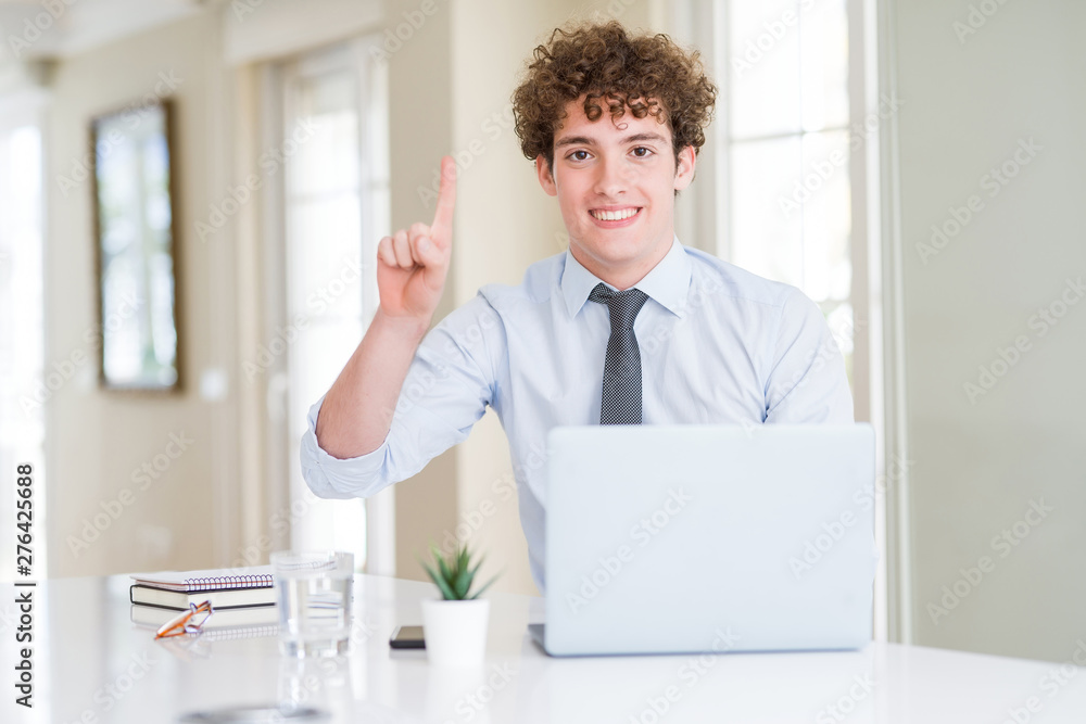 Young business man working with computer laptop at the office showing and pointing up with finger number one while smiling confident and happy.