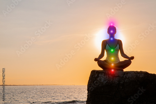 Woman is meditating with glowing seven chakras on stone at sunset. Silhouette of woman is practicing yoga on the beach.