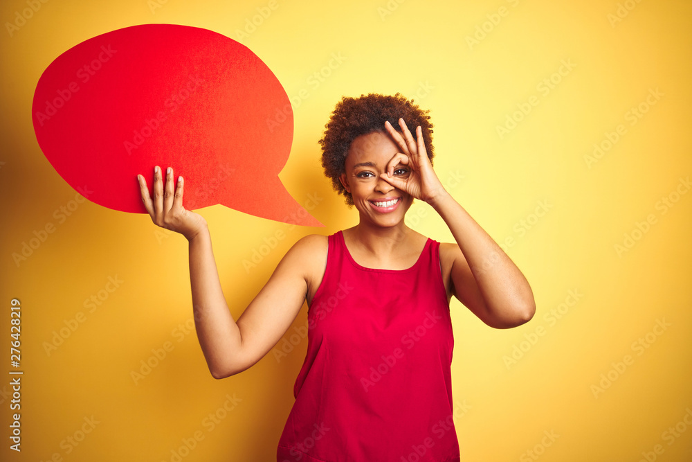Young african american woman holding speech bubble over yellow isolated background with happy face smiling doing ok sign with hand on eye looking through fingers Young african american woman holding speech bubble over yellow isolated background with happy face smiling doing ok sign with hand on eye looking through fingers