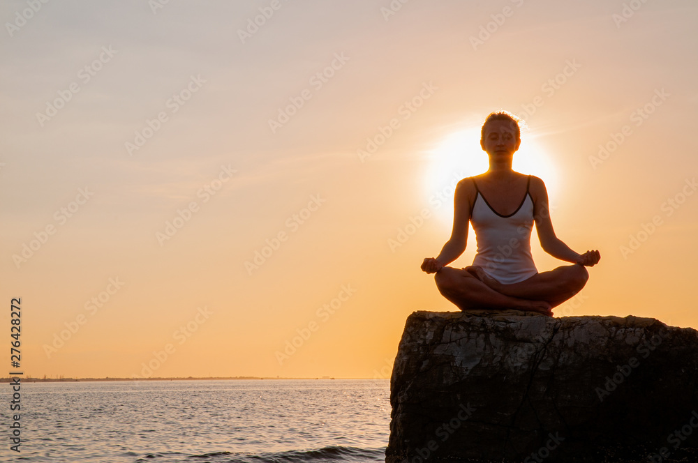 Woman is practicing yoga sitting on stone in Lotus pose at sunset. Silhouette of woman meditating on the beach