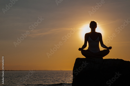 Woman is practicing yoga sitting on stone in Lotus pose at sunset. Silhouette of woman meditating on the beach