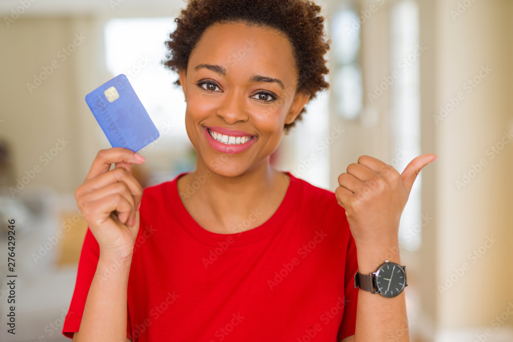 Young african american woman holding credit card pointing and showing with thumb up to the side with happy face smiling Young african american woman holding credit card pointing and showing with thumb up to the side with happy face smiling