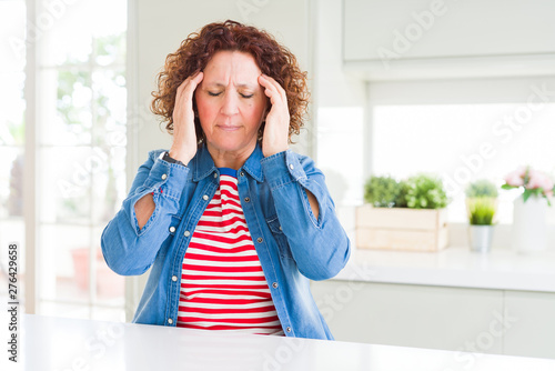 Middle age senior woman with curly hair wearing denim jacket at home with hand on headache because stress. Suffering migraine.