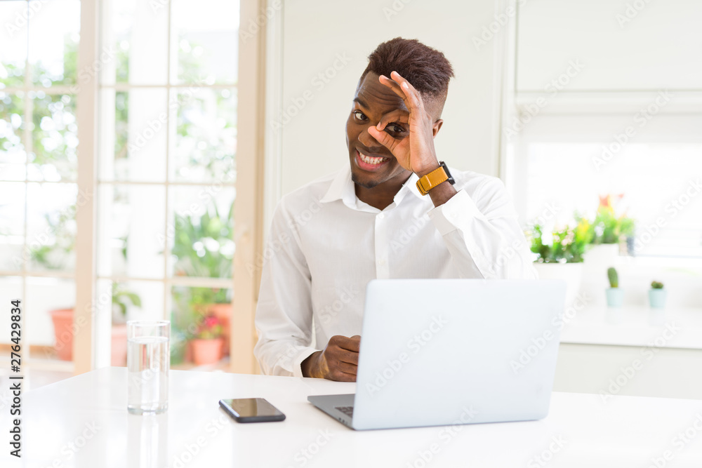 African american business man working using laptop doing ok gesture with hand smiling, eye looking through fingers with happy face.