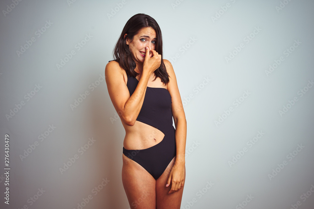 Beautiful woman wearing bikini swimwear over white isolated background smelling something stinky and disgusting, intolerable smell, holding breath with fingers on nose. Bad smells concept. Beautiful woman wearing bikini swimwear over white isolated background smelling something stinky and disgusting, intolerable smell, holding breath with fingers on nose. Bad smells concept.