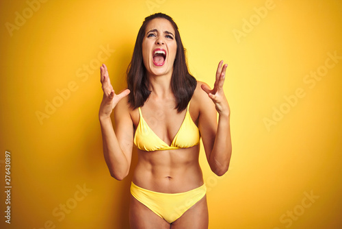 Beautiful woman wearing yellow bikini on summer over isolated yellow background crazy and mad shouting and yelling with aggressive expression and arms raised. Frustration concept.