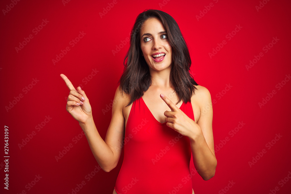 Beautiful woman wearing bikini swimwear over red isolated background smiling and looking at the camera pointing with two hands and fingers to the side. Beautiful woman wearing bikini swimwear over red isolated background smiling and looking at the camera pointing with two hands and fingers to the side.