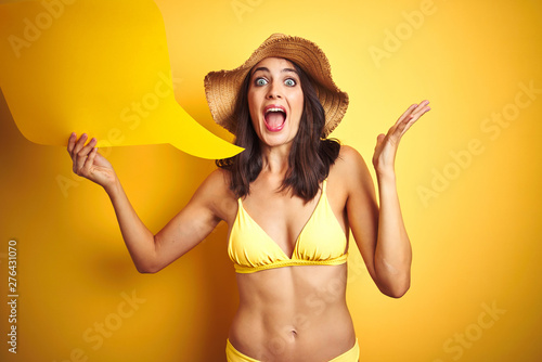 Beautiful woman wearing yellow bikini and holding talking balloon over isolated yellow background very happy and excited, winner expression celebrating victory screaming with big smile 
