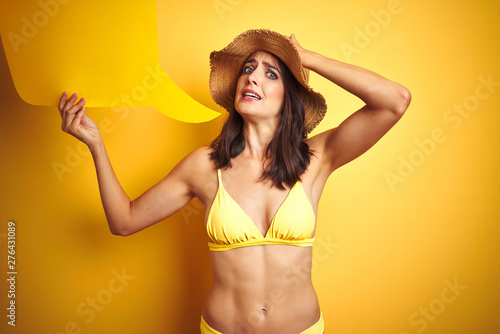 Beautiful woman wearing yellow bikini and holding talking balloon over isolated yellow background stressed with hand on head, shocked with shame and surprise face, angry and frustrated. 